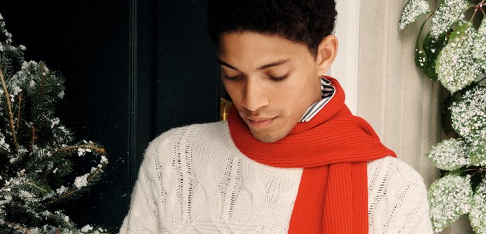Weisses Kabelstrickpullover, rot gerippter Schal, gestreifter Kragen. Schneebedeckter immergrüner Baum in der Nähe. Dunkle Tür im Hintergrund mit leichten Schneeverzierungen.
