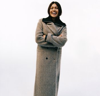 Langer grauer Wollmantel mit schwarzem Kragen, doppelreihigem Design, übergroßer Passform und sichtbaren Taschen. Model steht mit verschränkten Armen.