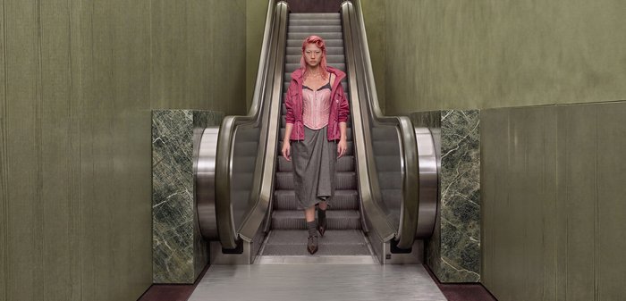 Une femme vêtue de rose descend un escalator dans un intérieur aux tons verts doux et aux murs revêtus de marbre.