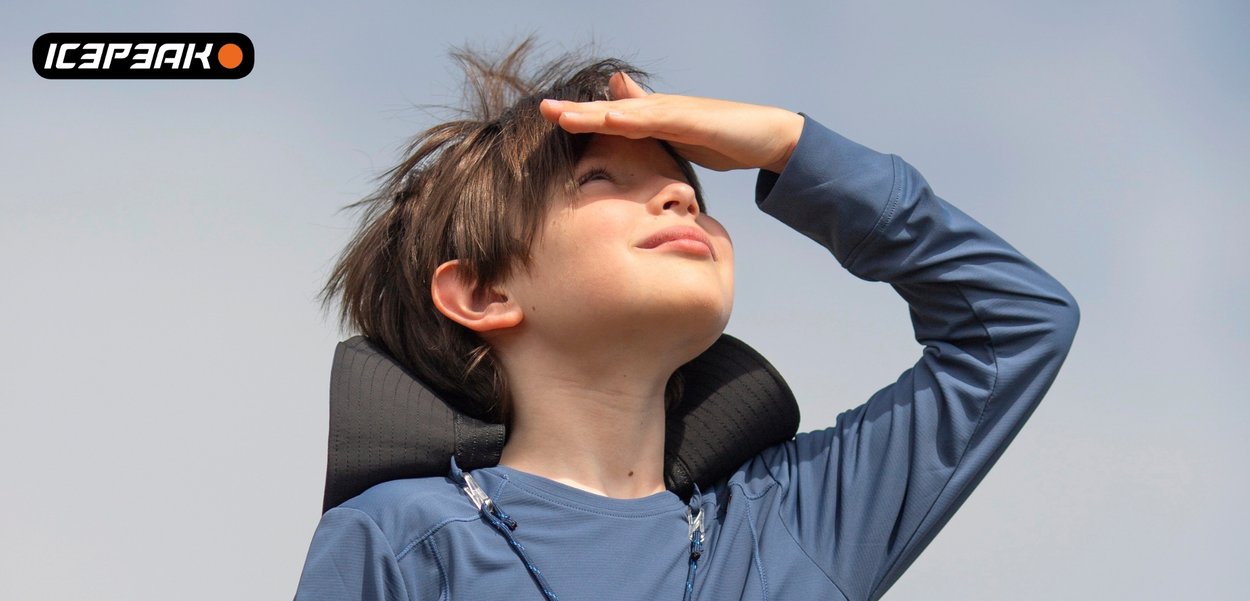 Kind trägt blaues Hemd und schwarzes Nackenkissen, hält sich die Augen zu und blickt zum hellen Himmel vor klarem Hintergrund.