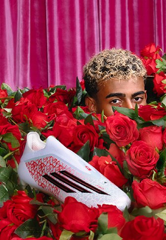 Personne aux cheveux blonds bouclés partiellement cachée parmi des roses rouges, avec une chaussure de football Adidas blanche et rouge au premier plan.