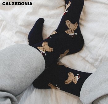 Pieds portant des chaussettes noires décorées d'oursons en bonnet de Noël, associés à un pantalon de survêtement gris sur une surface en tissu blanc.