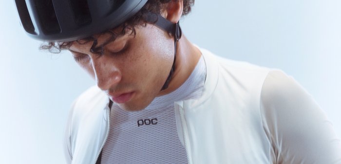 POC cycling jersey in light beige, short-sleeved with a textured white base layer underneath. Black helmet with air vents on head.