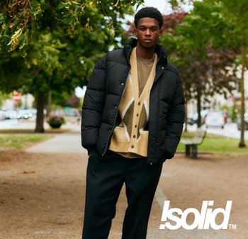 Schwarze Steppjacke über einer beigen Strickjacke mit Blattmuster, kombiniert mit weiten schwarzen Hosen. Außenaufnahme mit Grünpflanzen im Hintergrund.