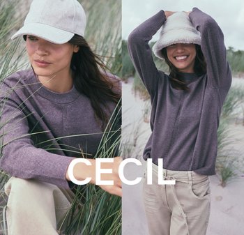 Lila Pullover mit strukturierter Oberfläche, beige Hose; das Model trägt eine helle Mütze und einen gemütlichen weißen Bucket Hat, vor einem grasbedeckten Strandhintergrund.