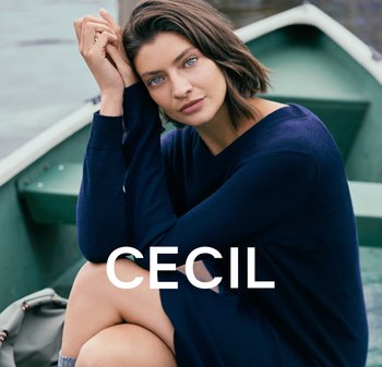Navyblauwe gebreide trui met V-hals en lange mouwen, met een zachte textuur. Model zittend op een boot, voor een deel gekruiste benen zichtbaar.