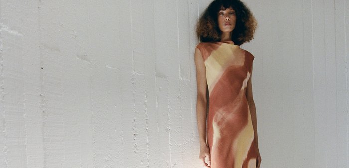 Femme aux cheveux bouclés se tenant sur une structure blanche incurvée, portant une longue robe sans manches avec des motifs abstraits orange et jaune et des sandales blanches.