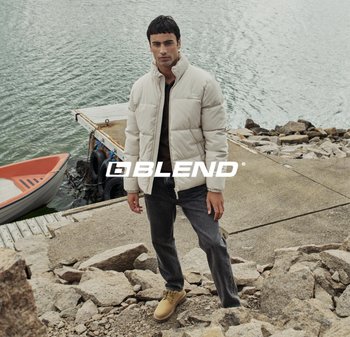 Beige pufferjack, grijze jeans en tan schoenen gedragen door een persoon die naast water staat. Rotsen en een boot zijn op de achtergrond.