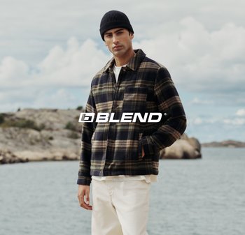 Camicia in flanella a quadri blu scuro con accenti verdi e beige, dotata di colletto, chiusura con bottoni e tasca frontale. Abbinata a pantaloni bianchi.