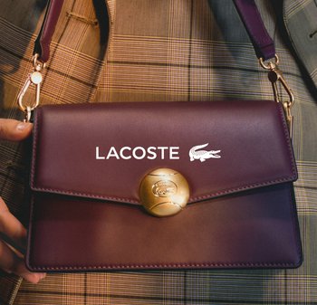 Bolso de cuero púrpura con un broche circular dorado, con el logo de Lacoste. Incluye una correa desmontable y textura suave.