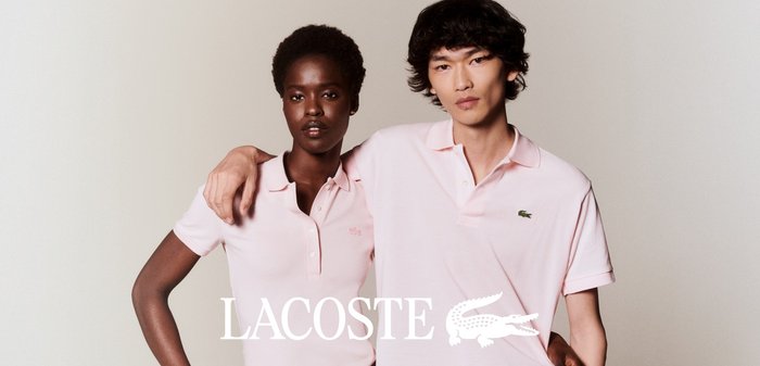 Deux mannequins portant des polos Lacoste rose clair sont proches, l'un avec le bras autour de l'épaule de l'autre sur un fond uni.