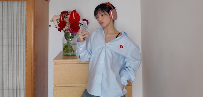 Mujer joven con una camisa azul de gran tamaño y auriculares rosas, mirando su teléfono inteligente, de pie junto a una cómoda de madera con un jarrón de flores.