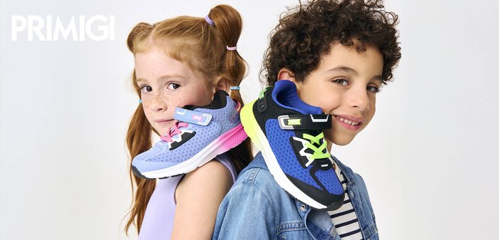Two children back-to-back, each holding a colorful sneaker on their shoulder; girl in purple, boy in denim jacket, white background.