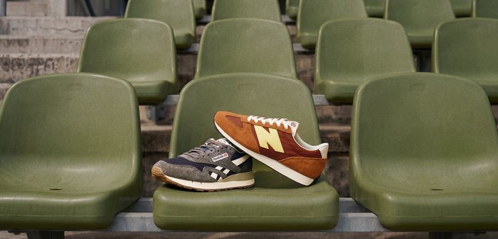 Deux baskets style vintage, une New Balance marron et une Reebok grise, placées sur des sièges verts en plastique dans un stade.