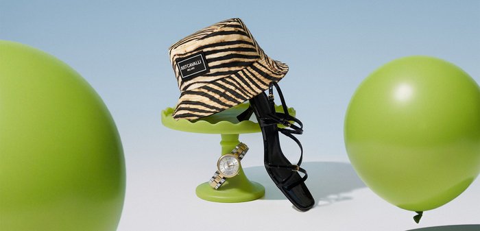 Chapeau seau rayé tigre, talon haut noir à lanières, et montre-bracelet en or exposés sur un piédestal vert entre deux ballons verts.