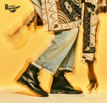 Botas de cuero negro con cordones y costuras amarillas, suela texturizada, combinadas con unos vaqueros de denim azul claro y una chaqueta con estampado frente a un fondo amarillo.