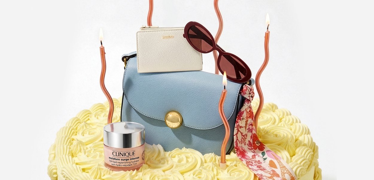 Bolso azul con cierre dorado, cartera blanca, gafas de sol rojas, pañuelo floral y crema Clinique sobre pastel amarillo glaseado con cuatro velas retorcidas encendidas.