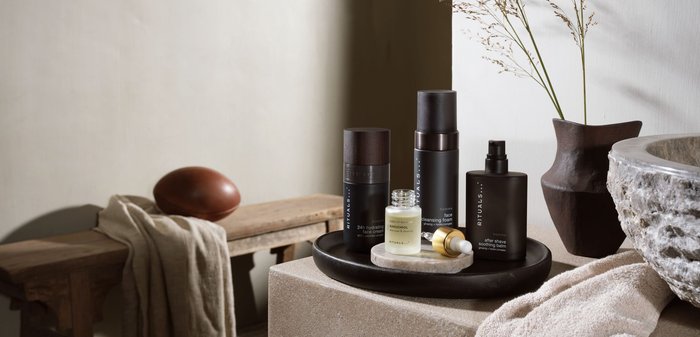 Un plateau noir contient quatre produits de soin de la peau : deux bouteilles foncées avec des bouchons en bois, un petit pot en verre et un tube. Surface en pierre beige et tissu doux.