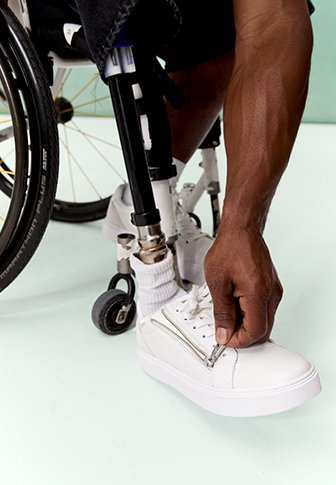 Person mit Beinprothese sitzt im Rollstuhl und schnürt einen weißen Sneaker mit seitlichem Reißverschluss auf hellem Boden.