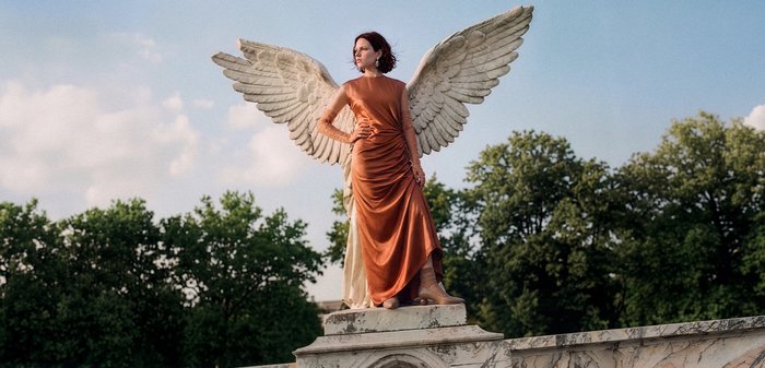 Femme en robe orange pose avec confiance sur une statue aux grandes ailes d’ange, entourée d’arbres sous un ciel bleu.