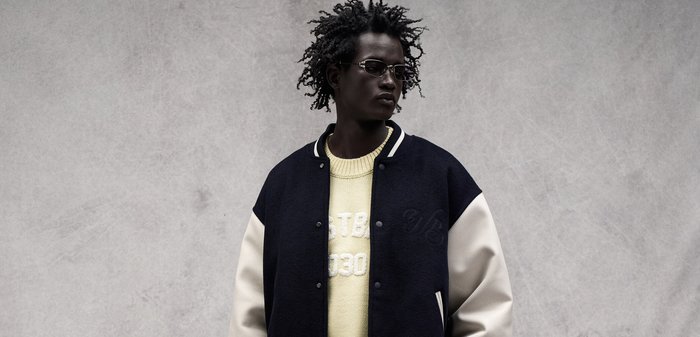 Uomo con capelli acconciati che indossa occhiali da sole, una giacca varsity bianca e nera e un maglione giallo chiaro su uno sfondo grigio uniforme.