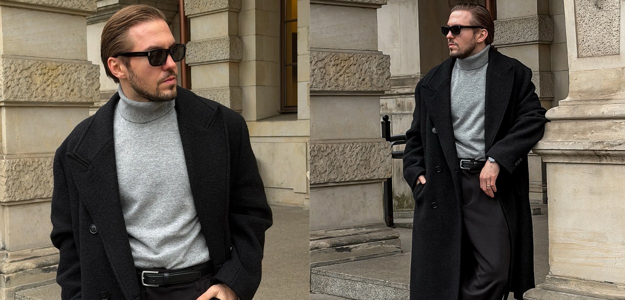 Bărbat purtând o haină neagră, pulover cu guler înalt gri, pantaloni negri și ochelari de soare, stând sprijinit de o clădire de piatră cu mâinile în buzunare.
