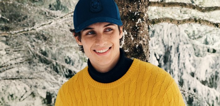 Gelbes Kabelstrickpullover mit hohem schwarzen Kragen, kombiniert mit einer blauen Mütze mit einem runden Logo, vor einem schneebedeckten Waldhintergrund.