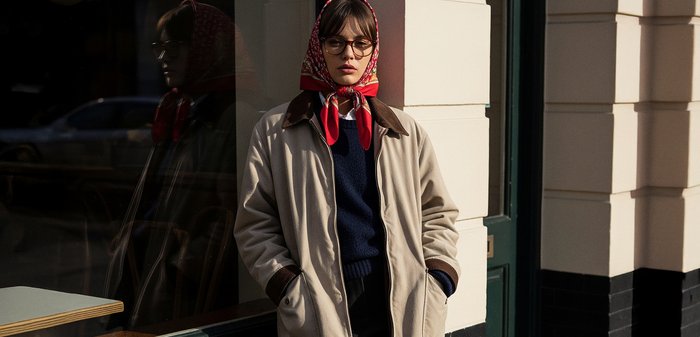 Jauna moteris dėvinti raudonos spalvos raštuotą galvos skarelę, akinius, smėlio spalvos paltą ir tamsiai mėlyną megztinį, stovinti prisidėjusi rankomis kišenėse prie pastato kampo su atspindinčiu stiklu.