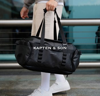 Černá sportovní taška s dvojitými uchy a ramenním popruhem. Vyrobená z hladkého, odolného materiálu. Na boku je logo a taška má zaoblený tvar.