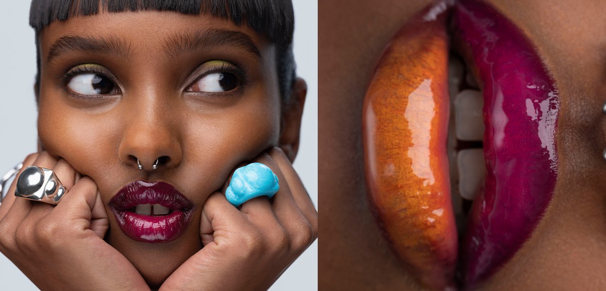 Osoba z kolczykiem w przegrodzie nosa, trzymająca twarz rękami z dużymi pierścionkami; zbliżenie na usta z połyskującą pomadką w kolorze pomarańczowym i magenty.