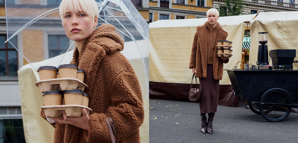 Brun, fluffig oversized kappa, ribbad brun kjol och knähöga bruna stövlar. Håller en bricka med kaffekoppar under ett genomskinligt paraply.