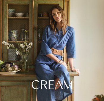 Woman in a blue dress with a woven belt sitting on a rustic wooden cabinet beside a vase with white flowers and a plate of artichokes.