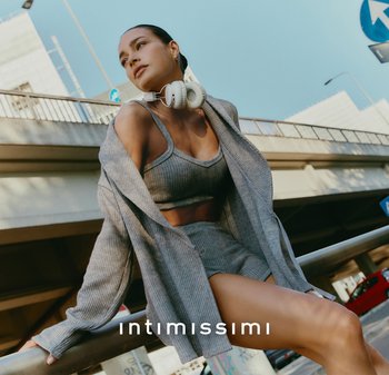 Set di top corto a costine grigio e pantaloni shorts abbinato a un lungo cardigan grigio, accessoriato con cuffie bianche. Sfondo urbano.