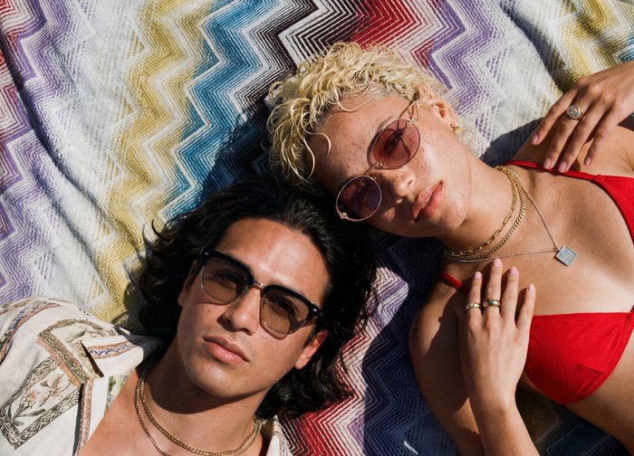 Δύο άτομα με γυαλιά ηλίου ξαπλωμένα σε ένα πολύχρωμο κουβέρτα με ζιγκ-ζαγκ σχέδιο· το ένα φοράει κόκκινο μπικίνι, το άλλο πουκάμισο με σχέδιο και χρυσές αλυσίδες.