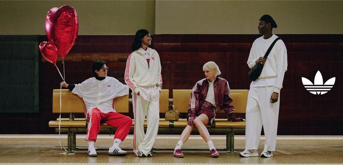 Woman in white and red tracksuit jacket sits inside a train, looking out of a window reflecting another woman in a maroon tracksuit jacket.