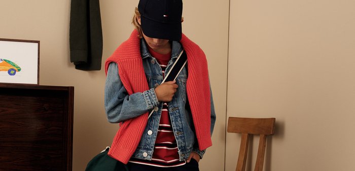 Kind, das eine Jeansjacke trägt, mit einem roten Pullover über die Schultern, einer blauen Mütze und einem gestreiften Hemd, das drinnen in der Nähe von Möbeln eine grüne Tasche trägt.