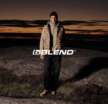 Uomo con felpa beige, gilet imbottito beige, pantaloni neri e stivali beige che sta in piedi su un terreno roccioso al tramonto con nuvole scure sopra.