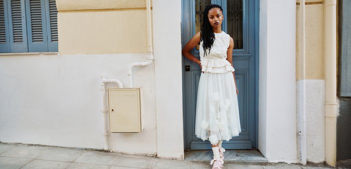Μια νέα γυναίκα με μακριά πλεξούδες φοράει κορυφή από λευκή δαντέλα και φούστα από τούλι, στέκεται σε μια πόρτα ενός φωτεινού κτιρίου με μπλε παντζούρια.