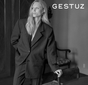 Crno-bijela slika koja prikazuje model u crnom blejzeru na prugice i odgovarajućim hlačama, s opuštenim krojem i strukturiranim dizajnom.