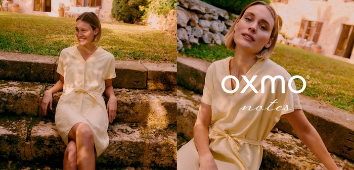 Junge Frau in einem hellgelben Kleid sitzt draußen auf Steinstufen, lächelnd, mit Grün und einem rustikalen Gebäude im Hintergrund.