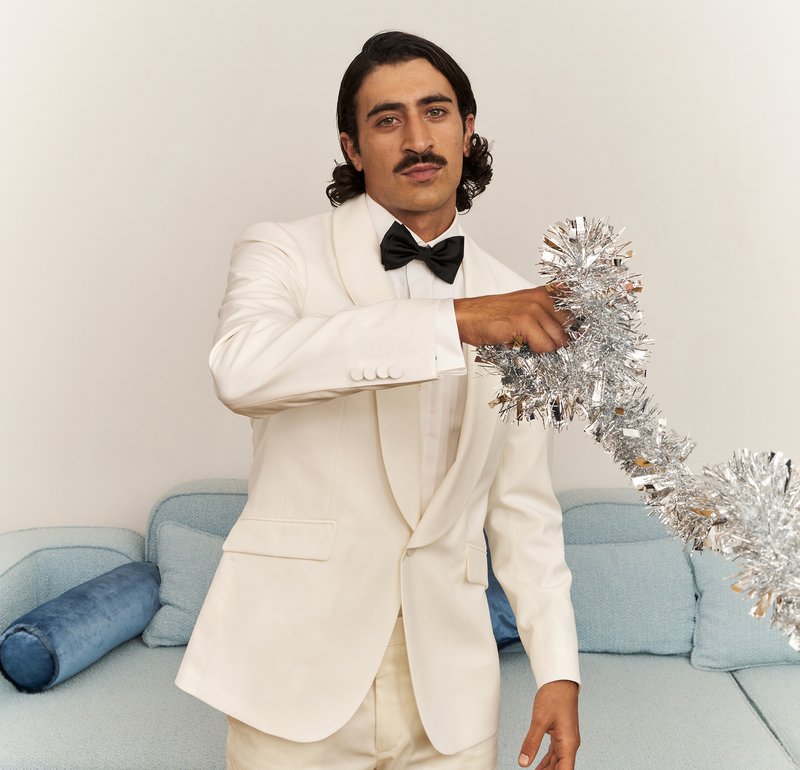 White tuxedo with a black bow tie, holding metallic silver tinsel, standing in a modern room with a light blue couch and cushions.