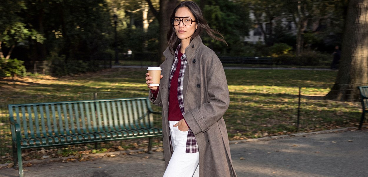 Una mujer con gafas y un abrigo gris pasea por un parque con un café para llevar en la mano, junto a un banco verde.