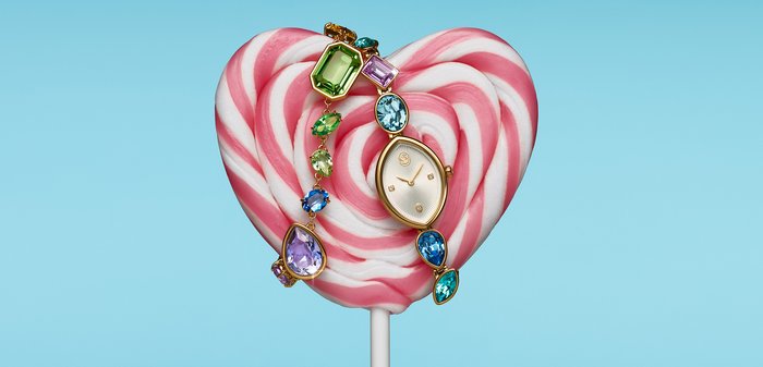 Montre en or avec des pierres précieuses multicolores drapée sur une sucette en forme de cœur rose et blanche sur un fond bleu clair.