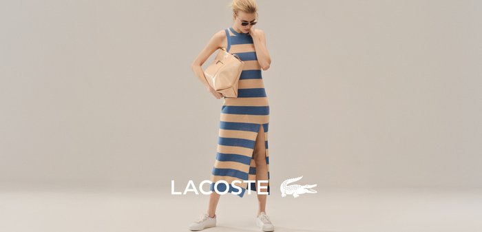 Mujer con vestido sin mangas a rayas azules y beige con abertura lateral, sosteniendo un bolso beige, usando gafas de sol; logotipo de Lacoste en la parte inferior.