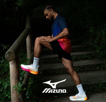 Las zapatillas deportivas Mizuno tienen una parte superior blanca con acentos en naranja y rosa, combinadas con pantalones cortos negros y una camiseta degradada de azul a rosa.