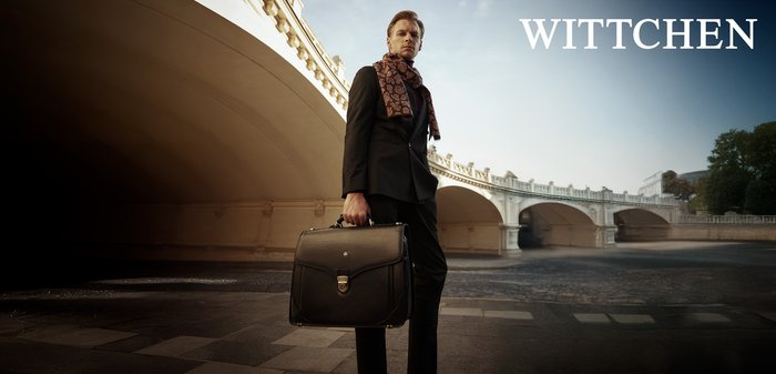 Czarna skórzana teczka z przednim klapkiem, srebrnymi akcesoriami i uchwytem. Tło przedstawia łukowy kamienny most oraz stonowane kolory.