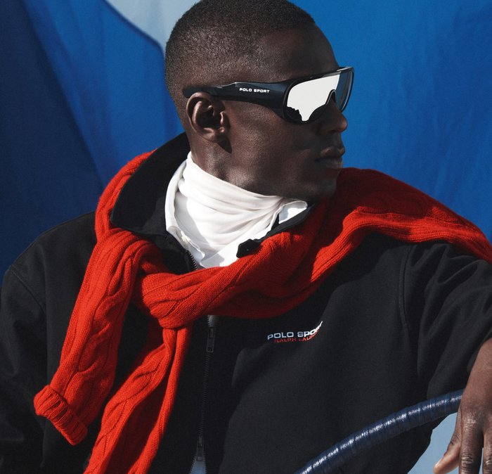 Hombre con gafas de sol Polo Sport reflectantes, chaqueta negra, camiseta de cuello alto blanca y suéter rojo sobre los hombros, sobre un fondo azul.
