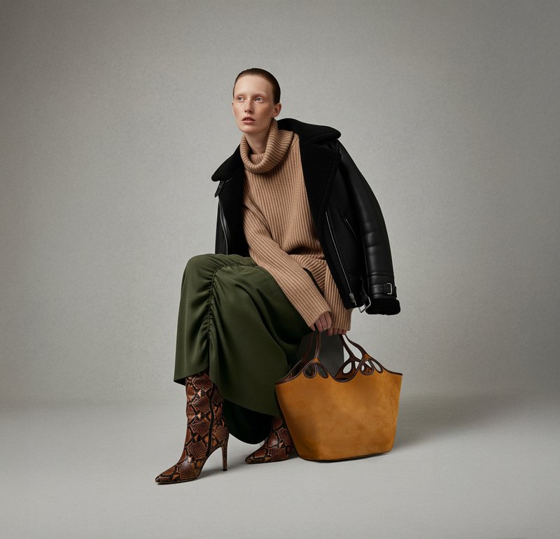 Tan ribbed sweater, black leather jacket, green skirt with ruching, snake-print boots, and a mustard yellow tote bag with cut-out handles.