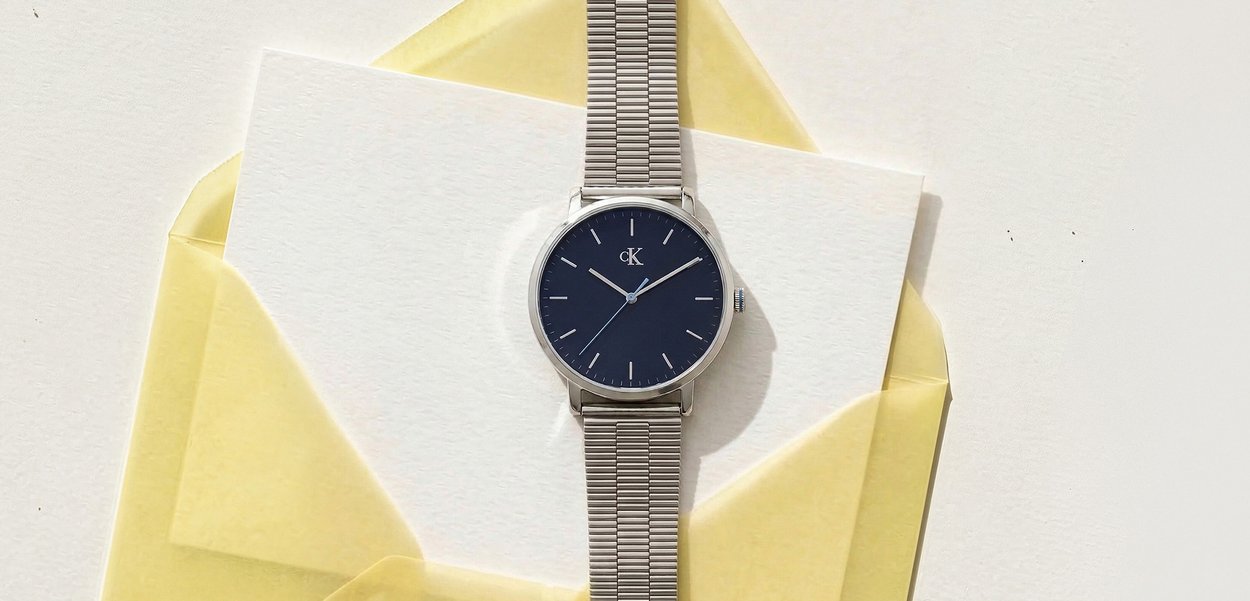 Reloj de pulsera Calvin Klein plateado con esfera negra y correa metálica apoyado sobre una tarjeta blanca dentro de un sobre amarillo.