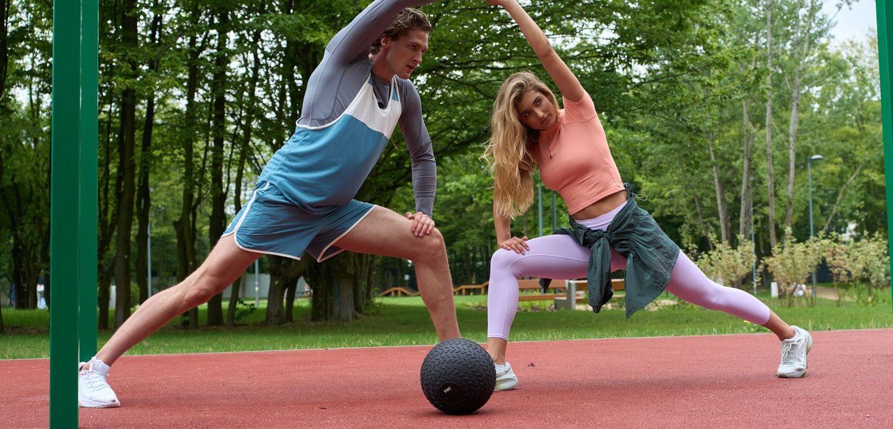 Dwie osoby rozciągają się na świeżym powietrzu na czerwonym gumowym podłożu. Mężczyzna ma na sobie jasnoniebieski i szary strój sportowy; kobieta nosi różową koszulkę i białe legginsy, z zieloną kurtką zawiązaną w talii. Pomiędzy nimi na ziemi leży teksturowana czarna piłka fitness.